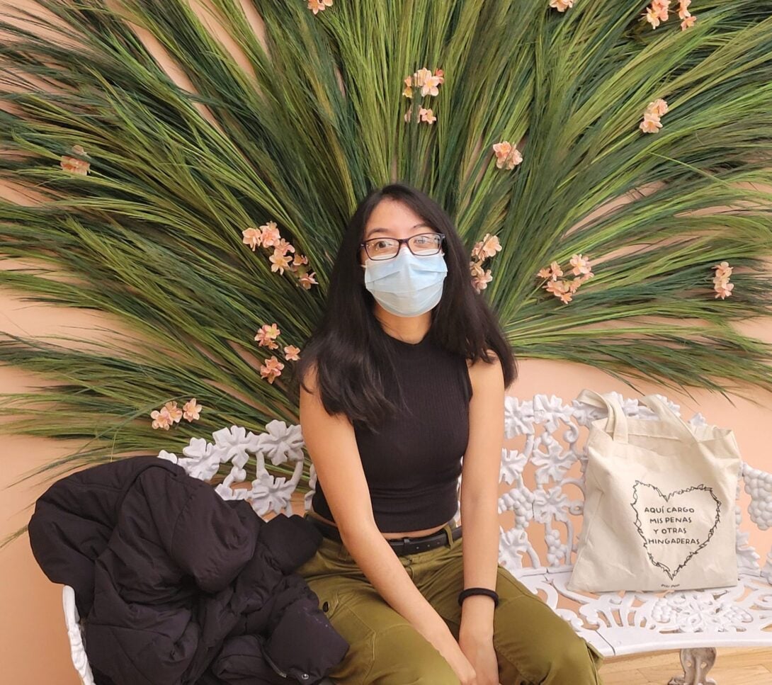 Light pink background decorated with green leaves and pink flowers. At the center, Cata Herrara sits on a white bench with her black coat and beige tote bag.