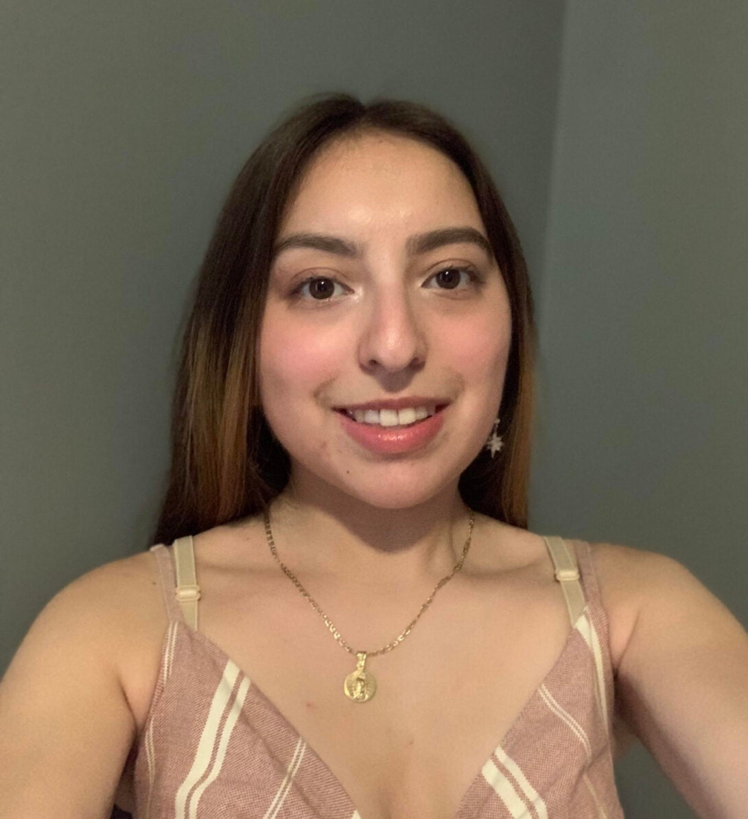 Dark grey background with Nayeli Jimenez in the center wearing a gold necklace and a striped pink and white top.