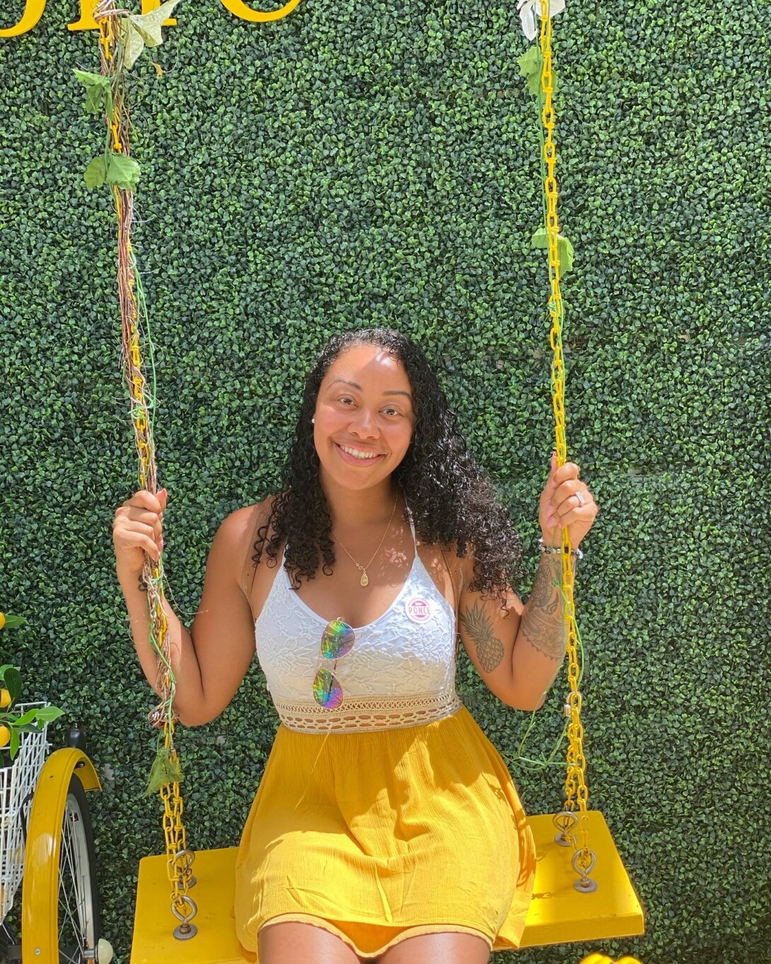 Green, grassy background, with Anissa Camacho, wearing a white tank top and a yellow skirt, is sitting on a yellow swing. The strings of the swing are also yellow and decorated with green leaves.