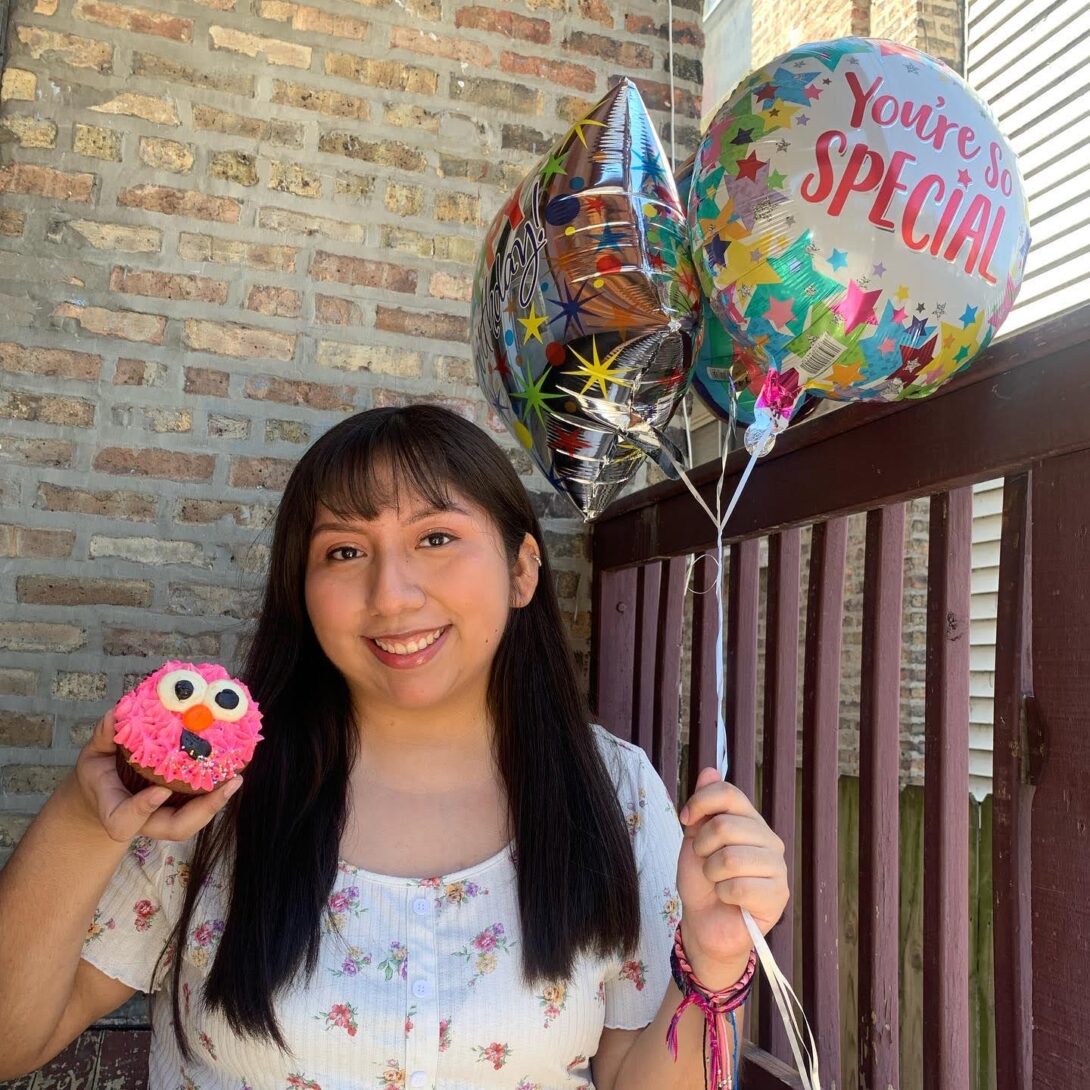 A light brown brick wall and dark cherry wooden fenced railing are in the background as Ana Martinez sits in the center. She is smiling and wearing a white shirt with pink flowers on it. Ana is also holding three balloons in one hand and a pink cupcake in the other.